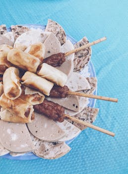 A plate of diverse Asian snacks including meat skewers and spring rolls on a blue textile.