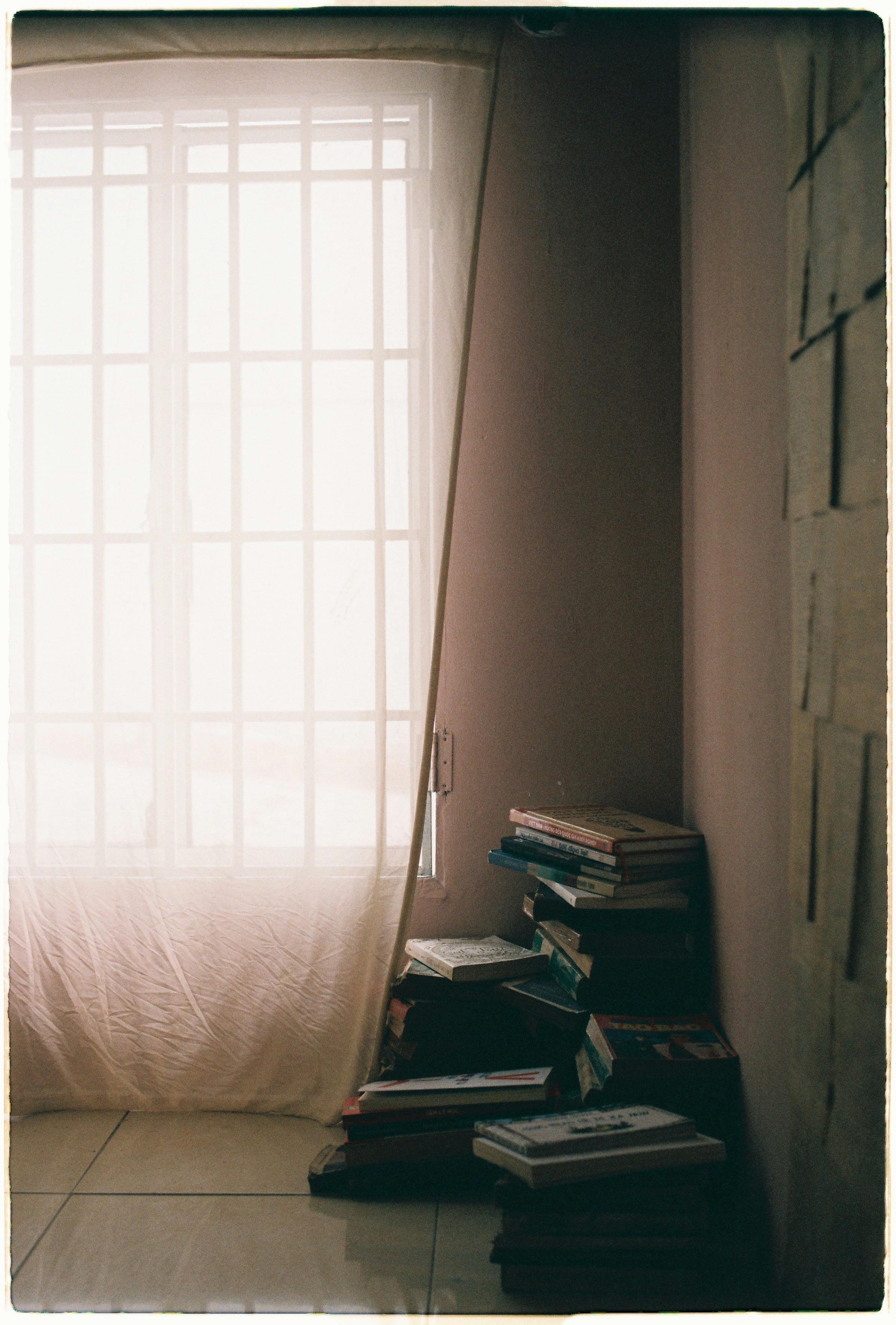 Free A stack of books by a softly lit window in a quiet, serene room. Stock Photo