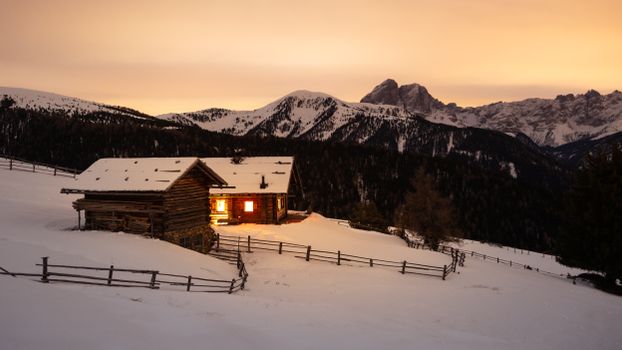 A wooden cabin glows warmly under a sunset amidst snowy Dolomites, creating a serene winter scene.