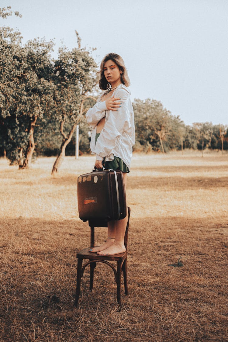 Woman Standing With Suitcase On Chair On Grassland