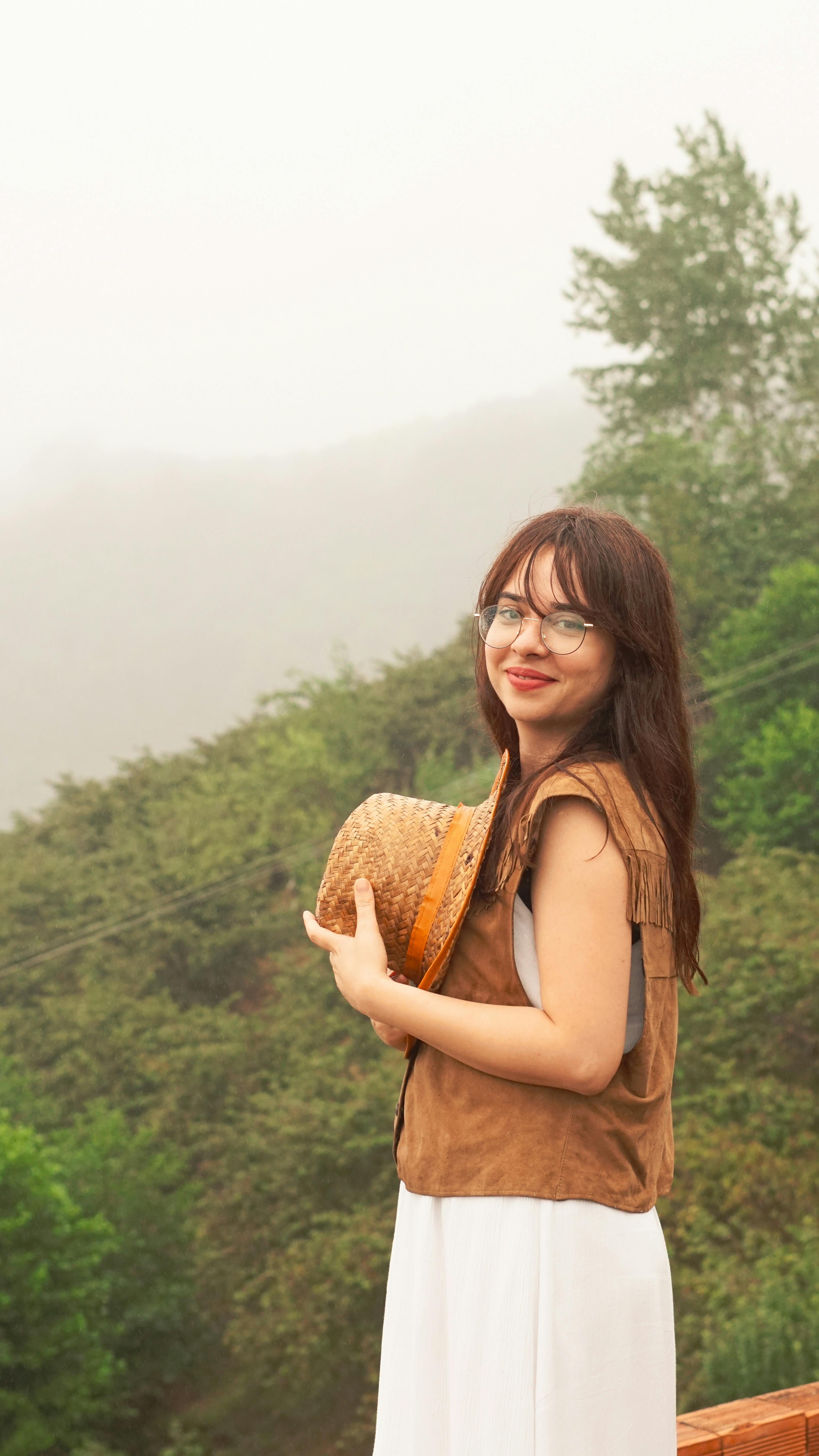 Smiling woman in nature holding a hat, enjoys a misty outdoor day.