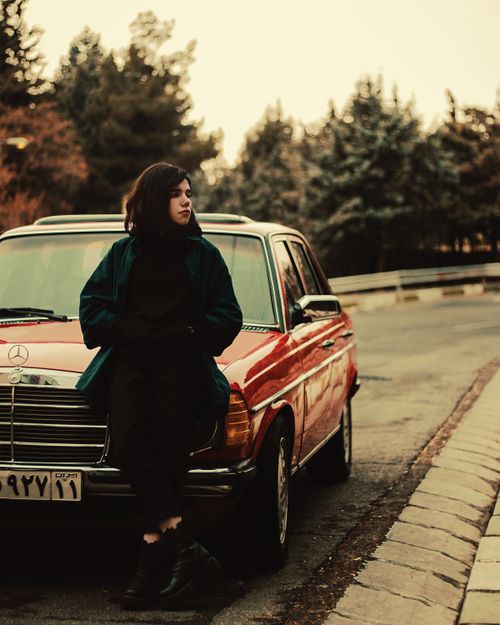 Free A woman leans on a vintage car on an urban street in Tehran, offering a nostalgic and moody vibe. Stock Photo