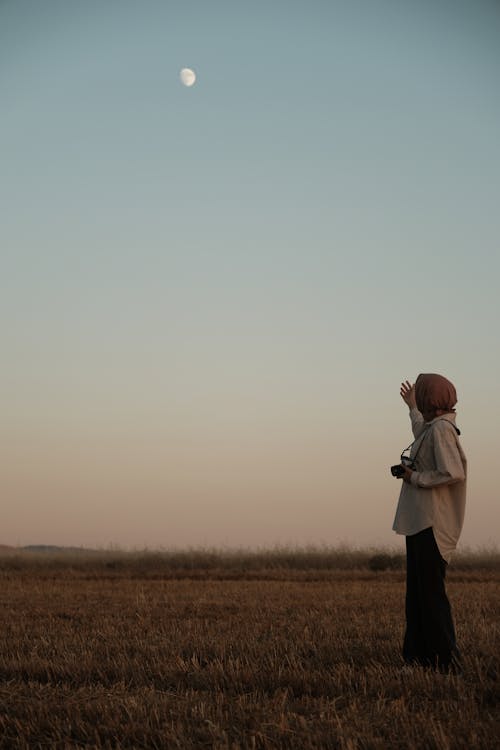 Woman in Hijab Standing on Rural Field with Moon on Clear Sky