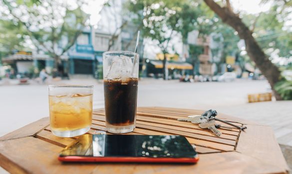 Glasses of iced drinks with smartphone on wooden table in a lively outdoor cafe setting.
