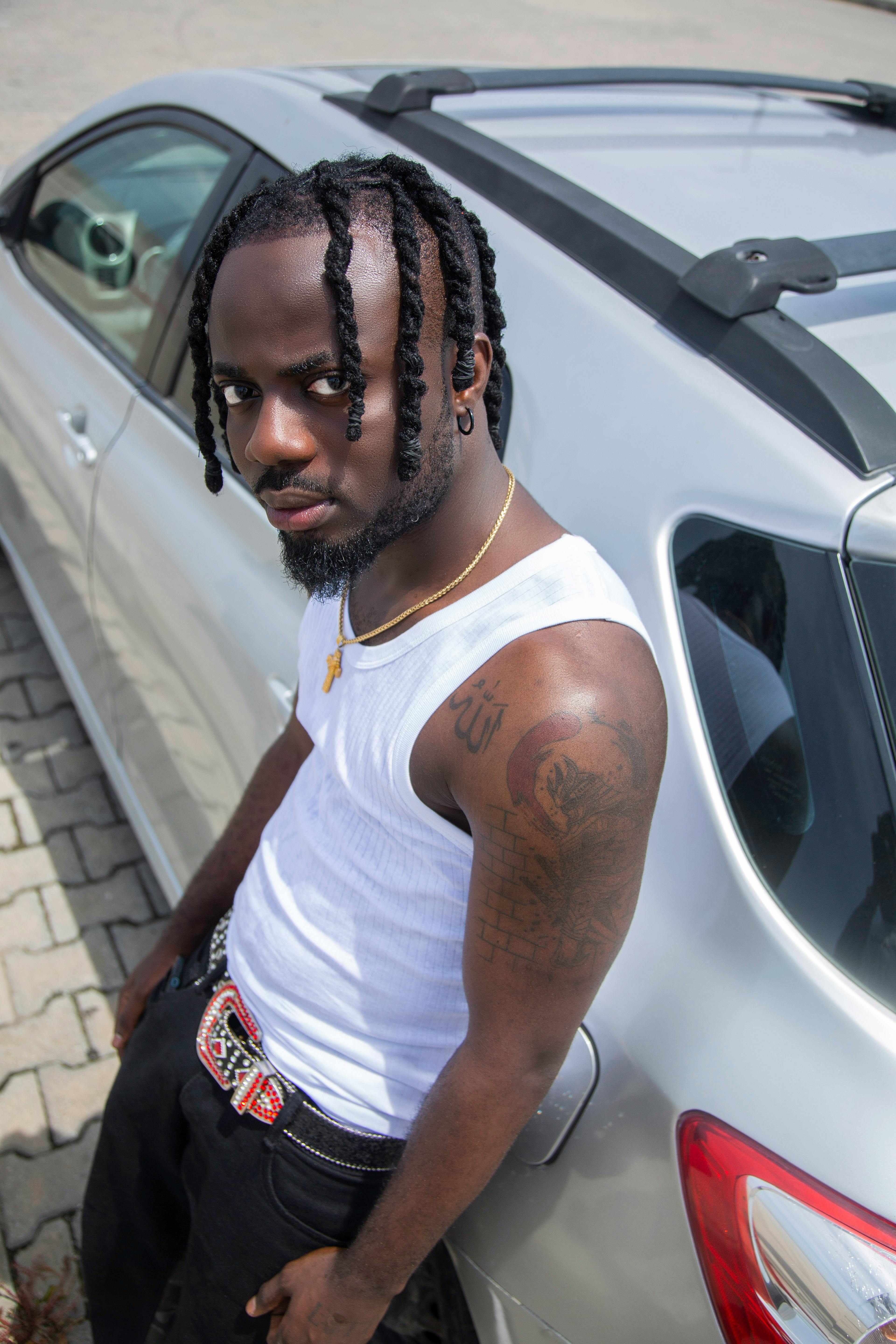Free Young man with braids and tattoos leaning on a car in a parking lot. Stock Photo