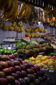 A colorful array of tropical fruits including mangoes, bananas, and pineapples at an indoor market stall.