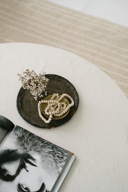 A minimalist still life depicting a necklace, dried flowers, and a book on a table, captured in soft lighting.