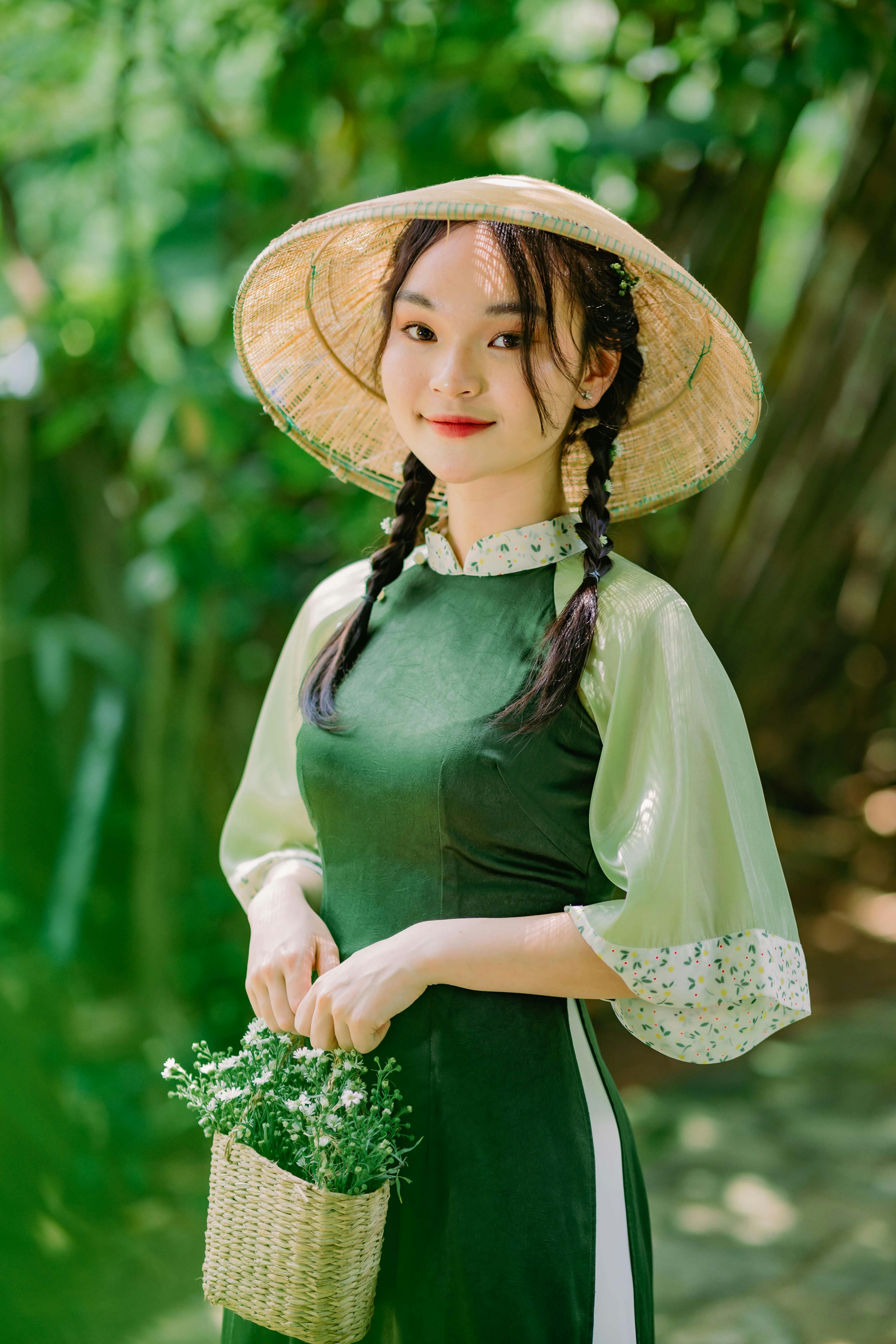 Portrait of an Asian woman in a green dress and conical hat holding a basket, surrounded by lush nature.