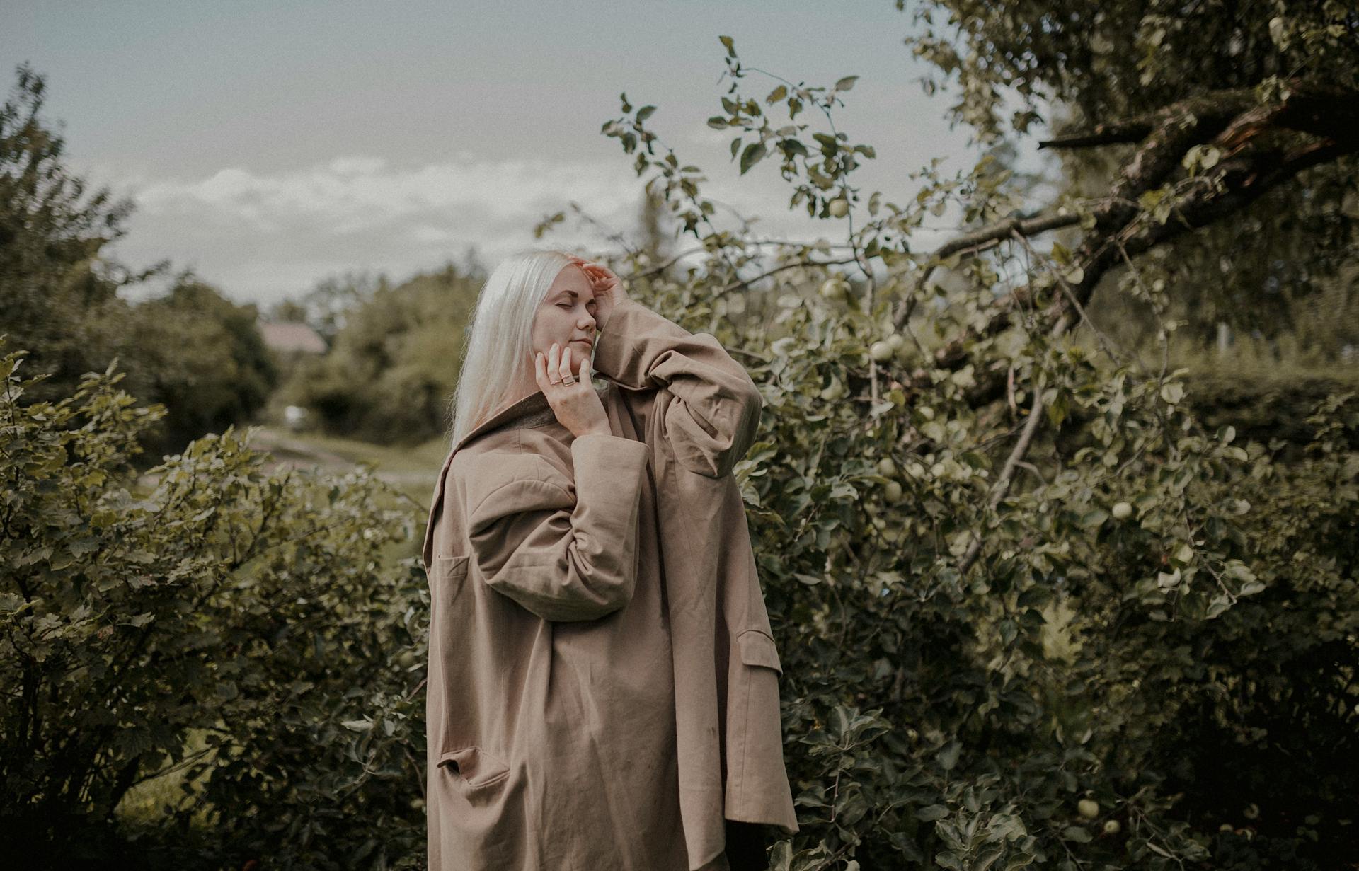 Elegant woman with platinum blond hair in a coat posing with eyes closed outdoors in summer.