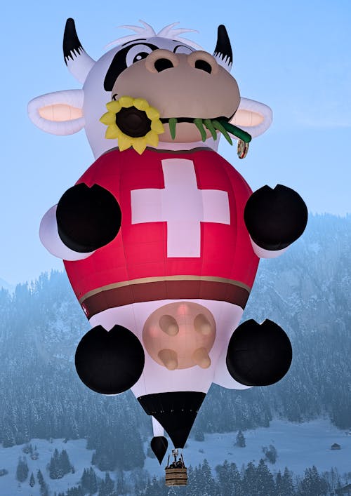 A Swiss Cow Bluemli Hot Air Balloon Flying over a Snowy Valley 