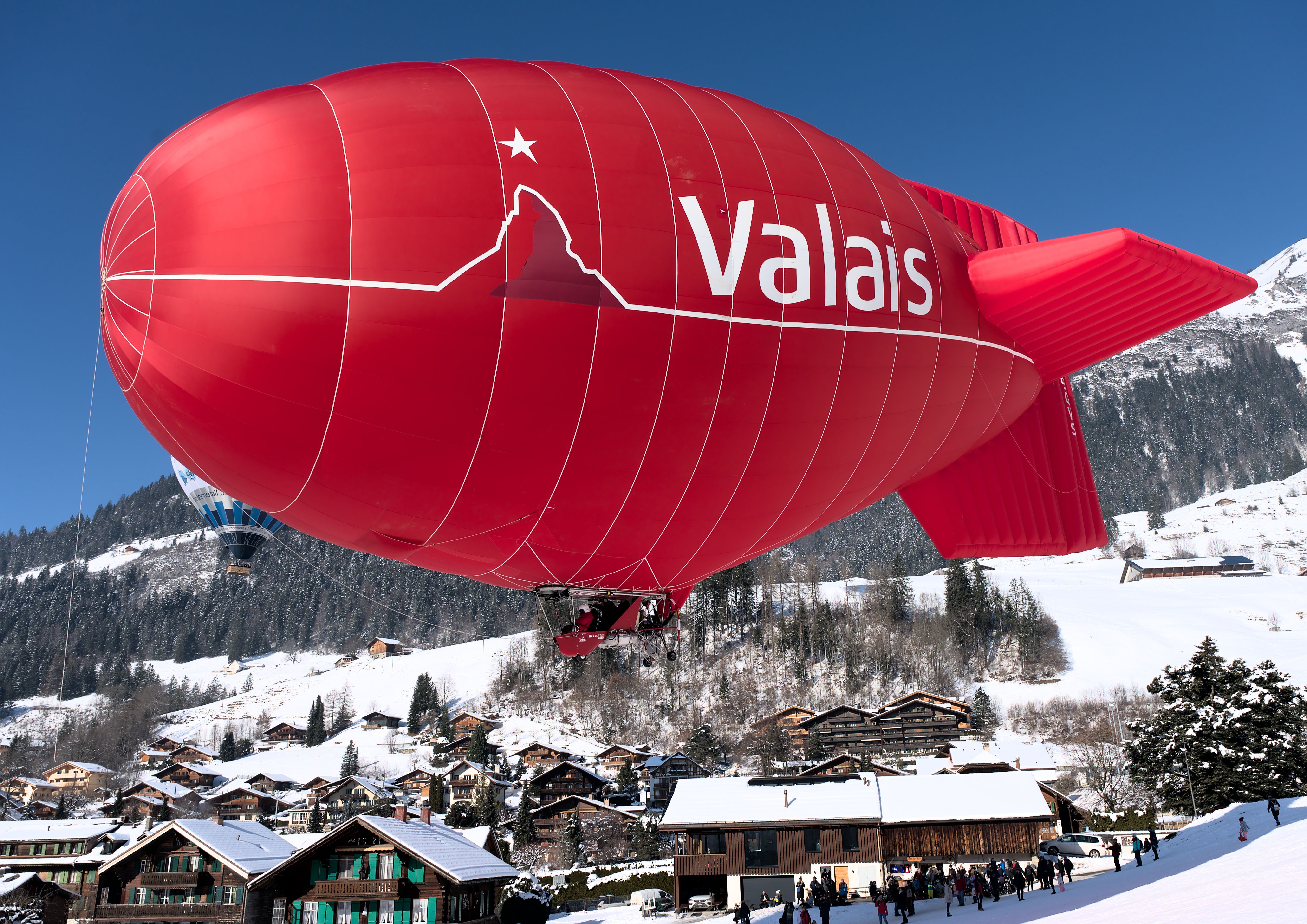 A vibrant red blimp flies over a snowy village in Château-d’Œx, Switzerland, showcasing winter adventure.