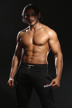Muscular male model with long hair posing topless against a black background.