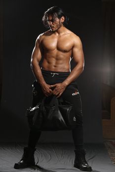 Shirtless muscular man holding a leather sports bag in a studio setting.