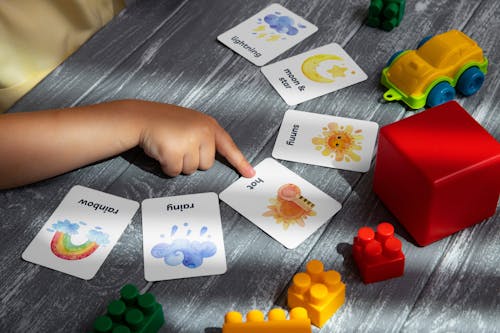 Close-up of a Child Pointing at a Card Lying on a Table with Toys 