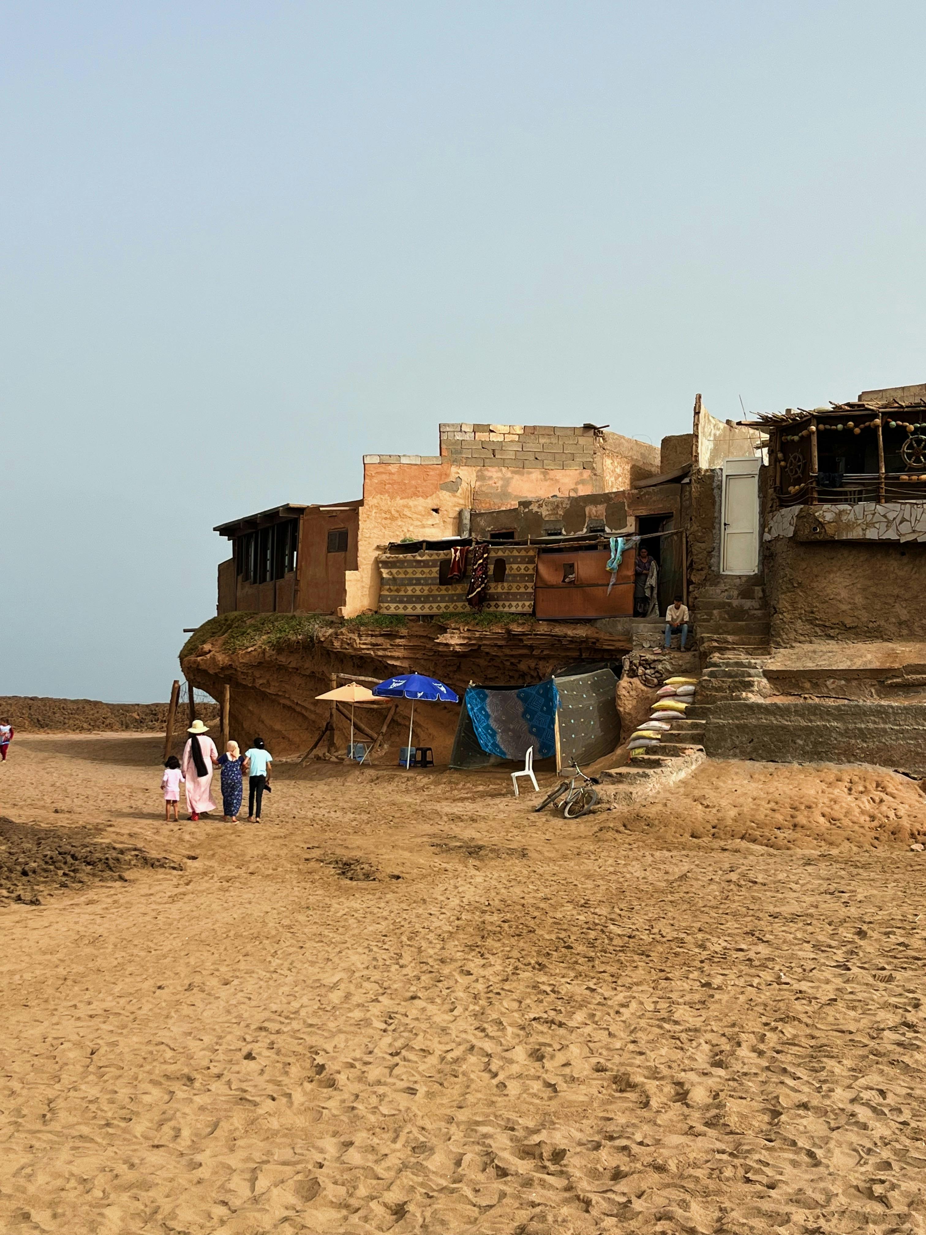 A rural village scene showing old buildings and people walking on sandy terrain under clear skies.