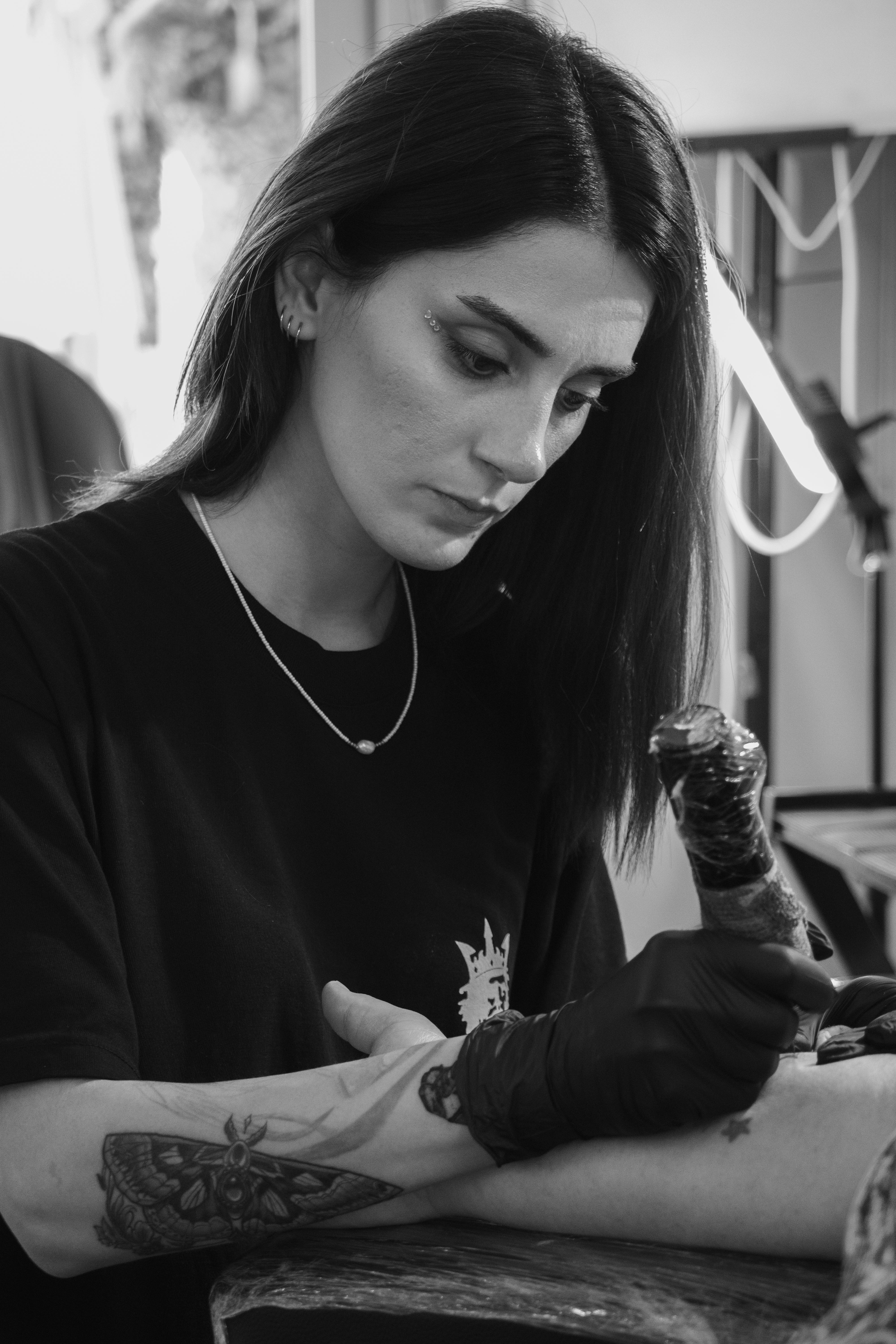A skilled female tattoo artist works intently on a client's arm in black and white.