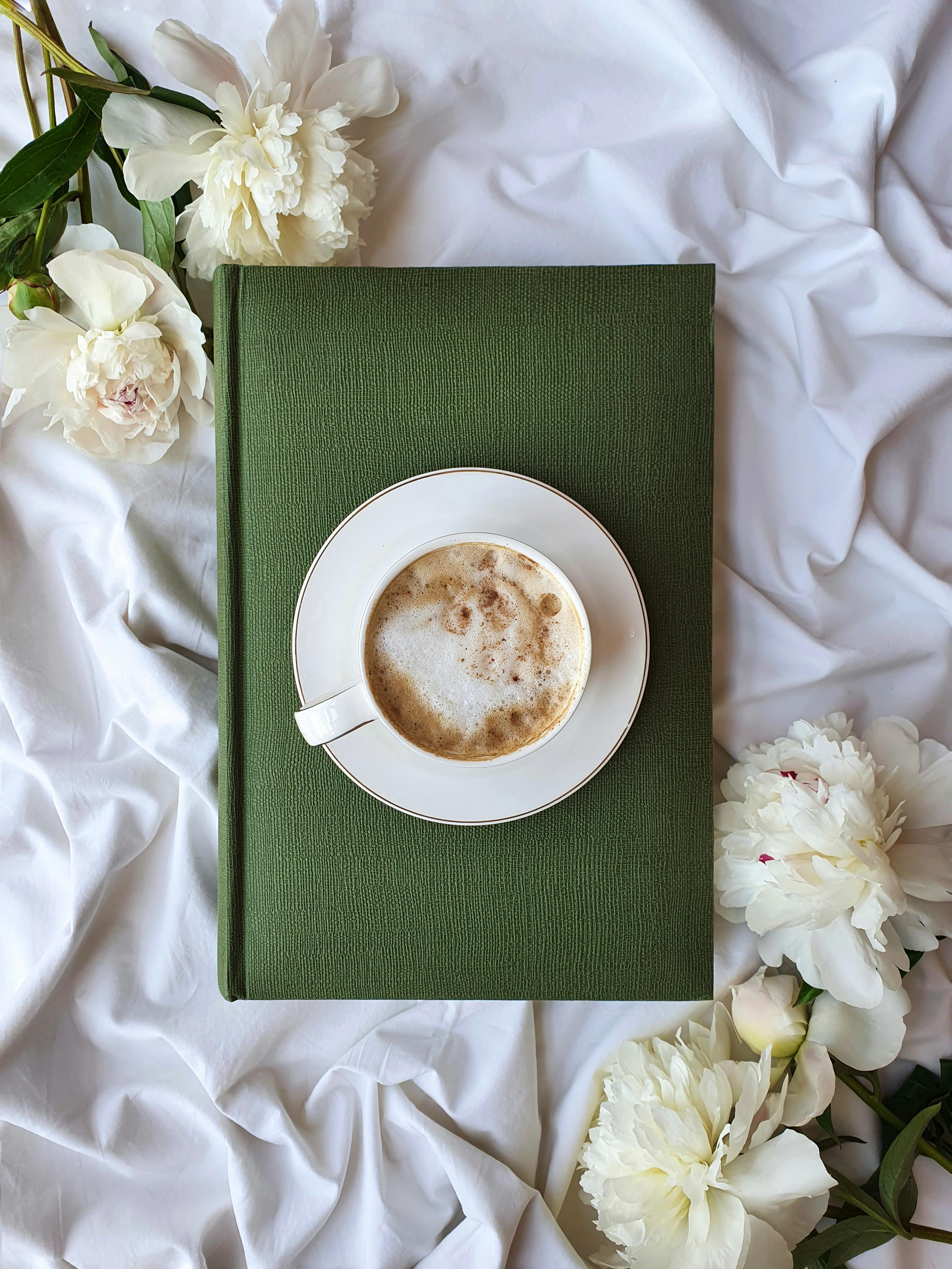 Top view of a coffee cup on a green book surrounded by white flowers, creating a serene vibe.