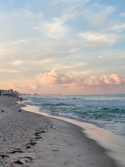Free Peaceful beach at sunset featuring ocean waves and distant skyscrapers under a pastel sky. Stock Photo