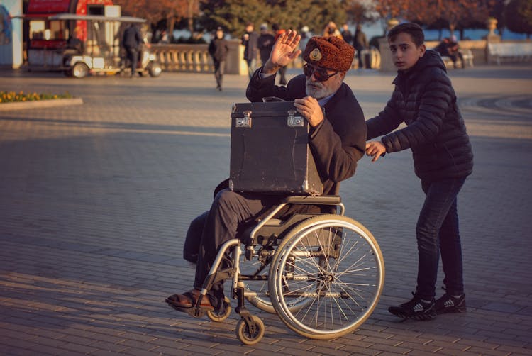 A Boy Pushing A Wheelchair With An Elderly Man