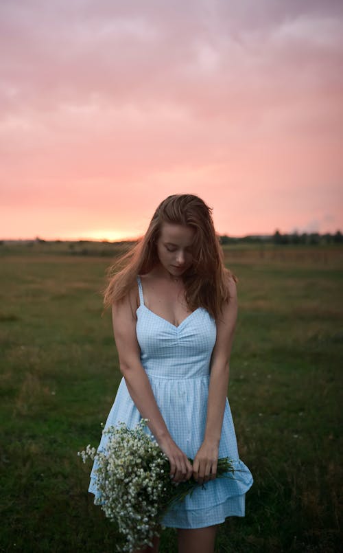 Young Woman in a White Dress Standing on a Meadow and Holding a Bunch of Flowers