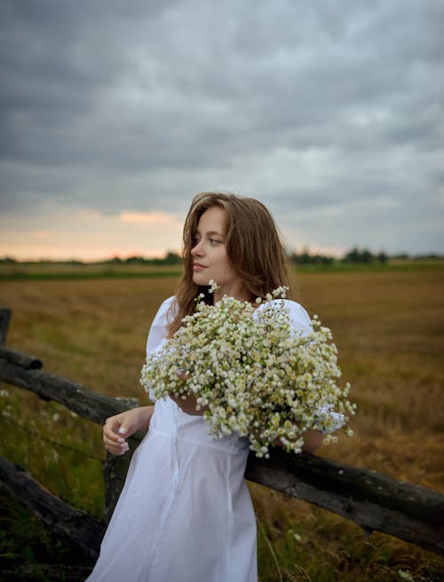 Young Woman in a White Dress Standing on a Meadow and Holding a Bunch of Flowers