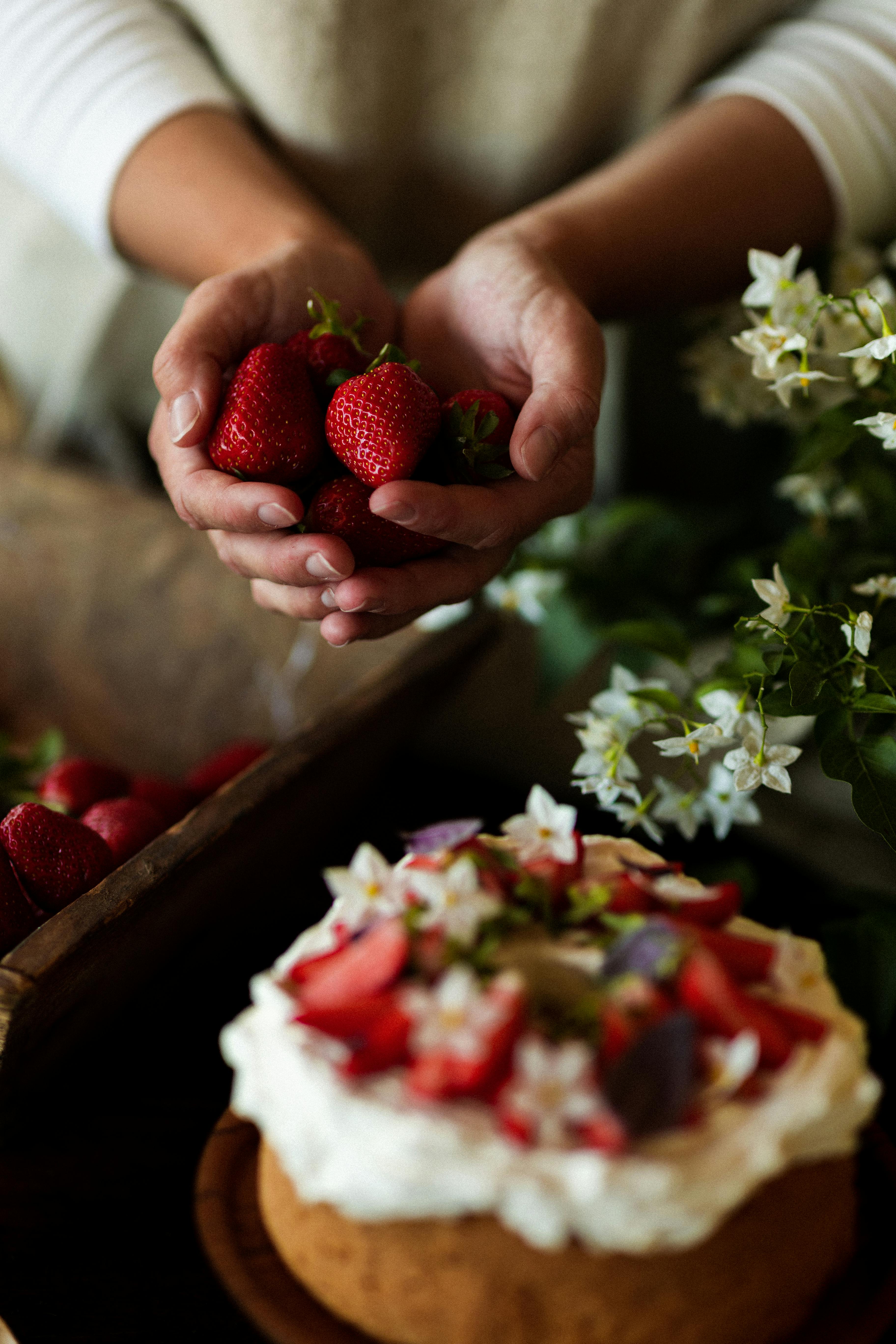 Close-up of hands holding strawberries over a cream-topped cake with flowers, showcasing fresh ingredients.