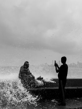 A man photographs waves splashing on a seaside promenade, capturing the moment on a smartphone.