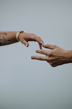 A touching moment depicted by two hands reaching out, featuring tattoos and a watch, set against a neutral background.