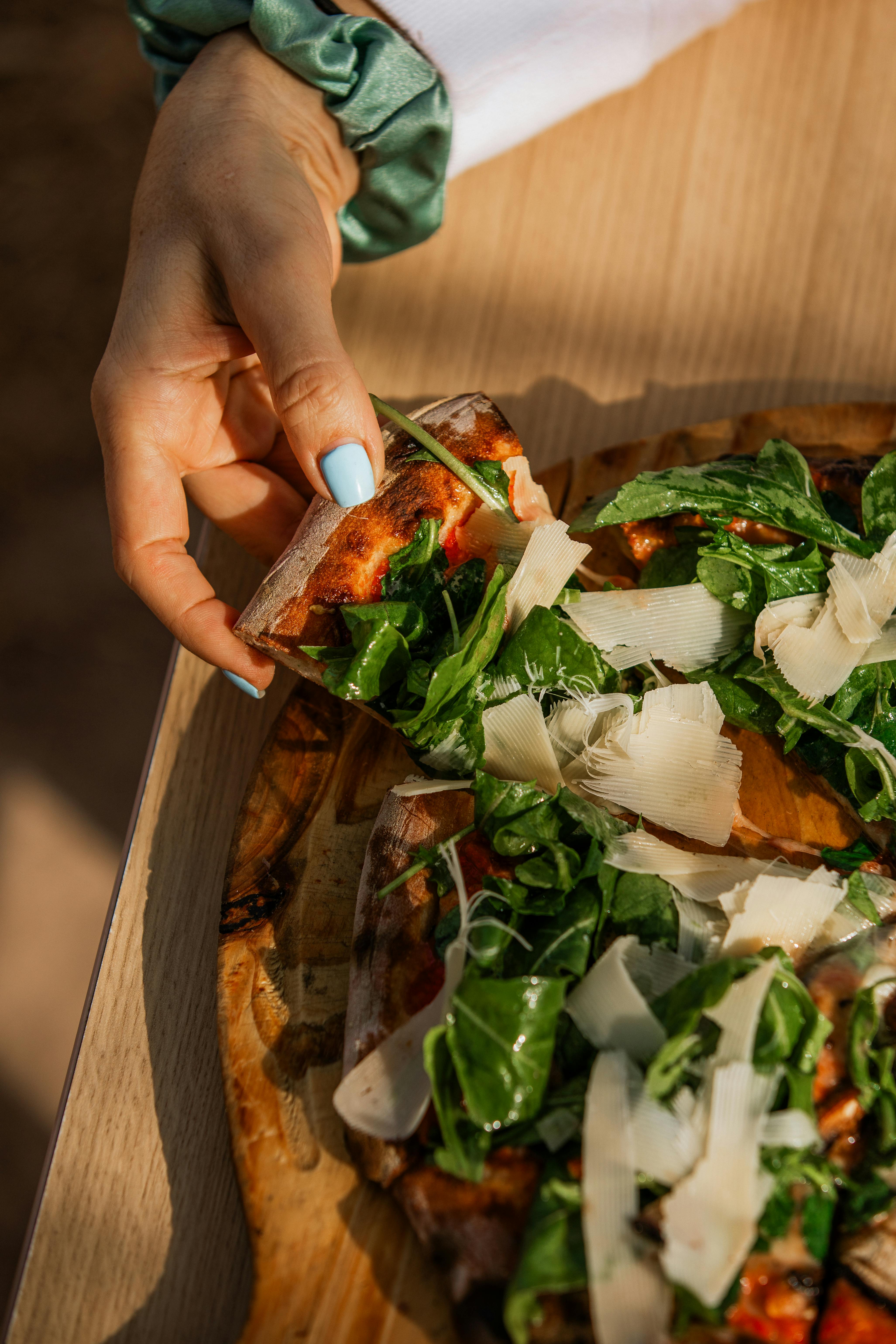 A close-up of a hand with manicured nails holding a slice of pizza topped with fresh greens and cheese.