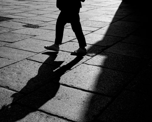 Free Black and white image of a pedestrian walking with a distinct shadow on pavement. Stock Photo
