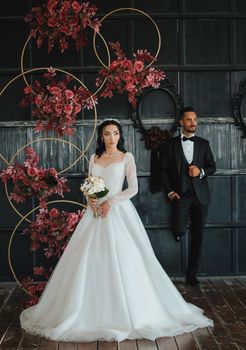A stylish couple in wedding attire poses with a floral backdrop, showcasing elegance and romance.