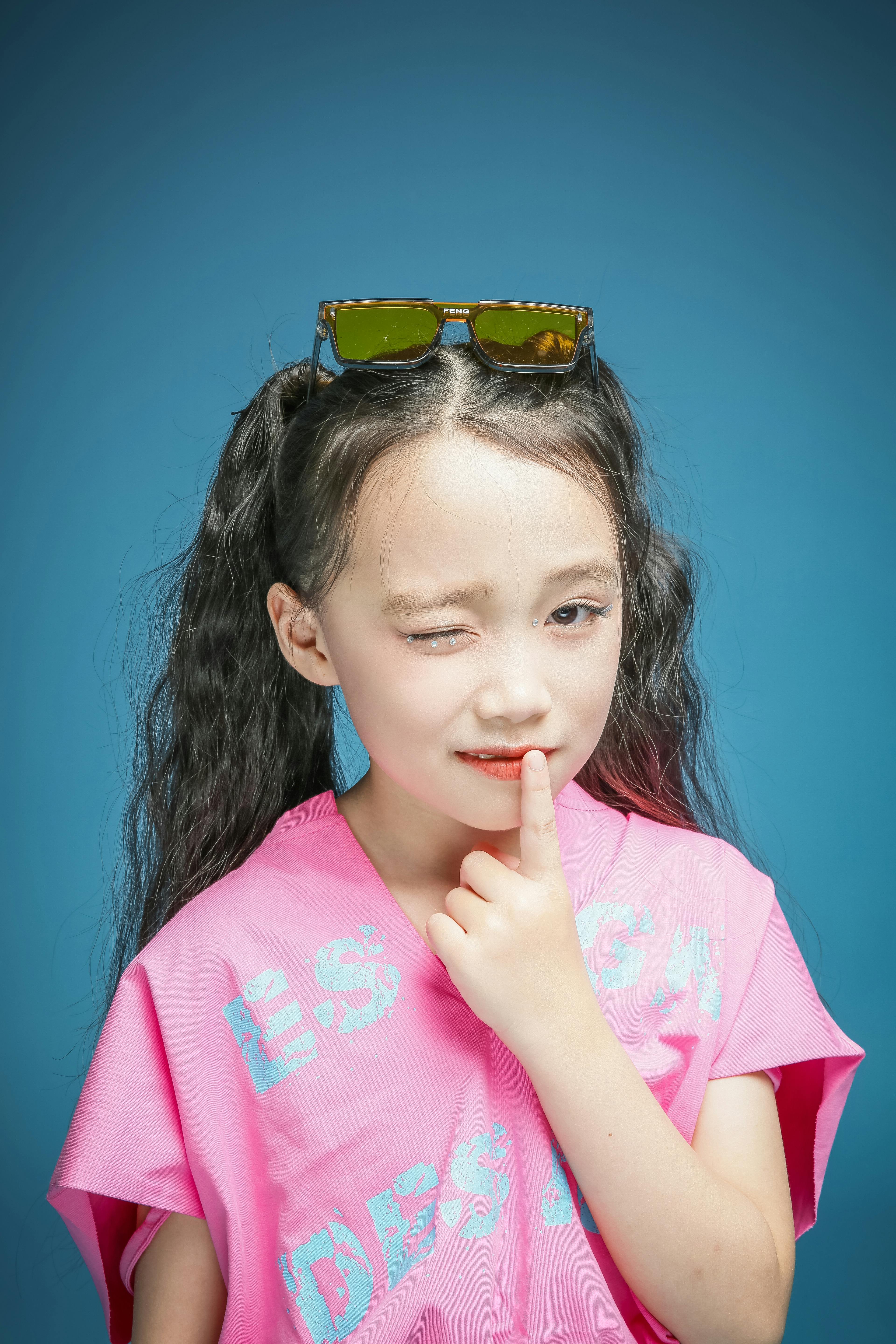 A young girl poses confidently in a pink t-shirt and sunglasses against a blue background.