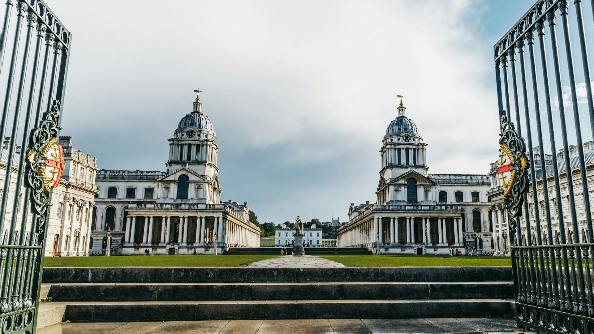 The iconic domes of the Royal Naval College in Greenwich, England framed by ornate gates, under a bright sky.