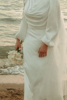Elegant bride in a white gown holding a flower bouquet by the beach. Romantic and serene setting.