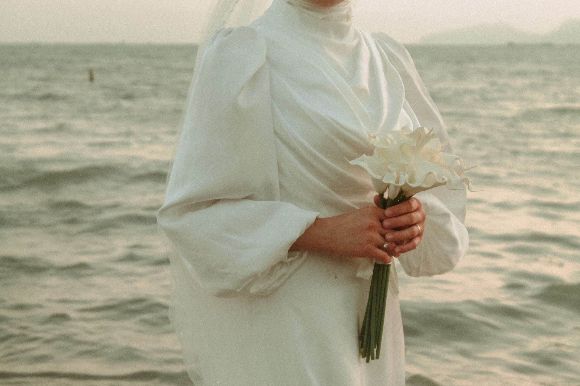 A bride in a white gown holds calla lilies by the beach, exuding elegance and serenity.