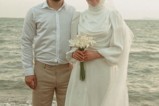 A newlywed couple posing in elegant attire by the sea, capturing a romantic beach wedding moment.