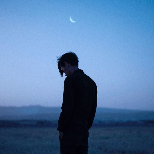 Serene silhouette of a man standing in a field under a crescent moon during twilight with a peaceful sky.