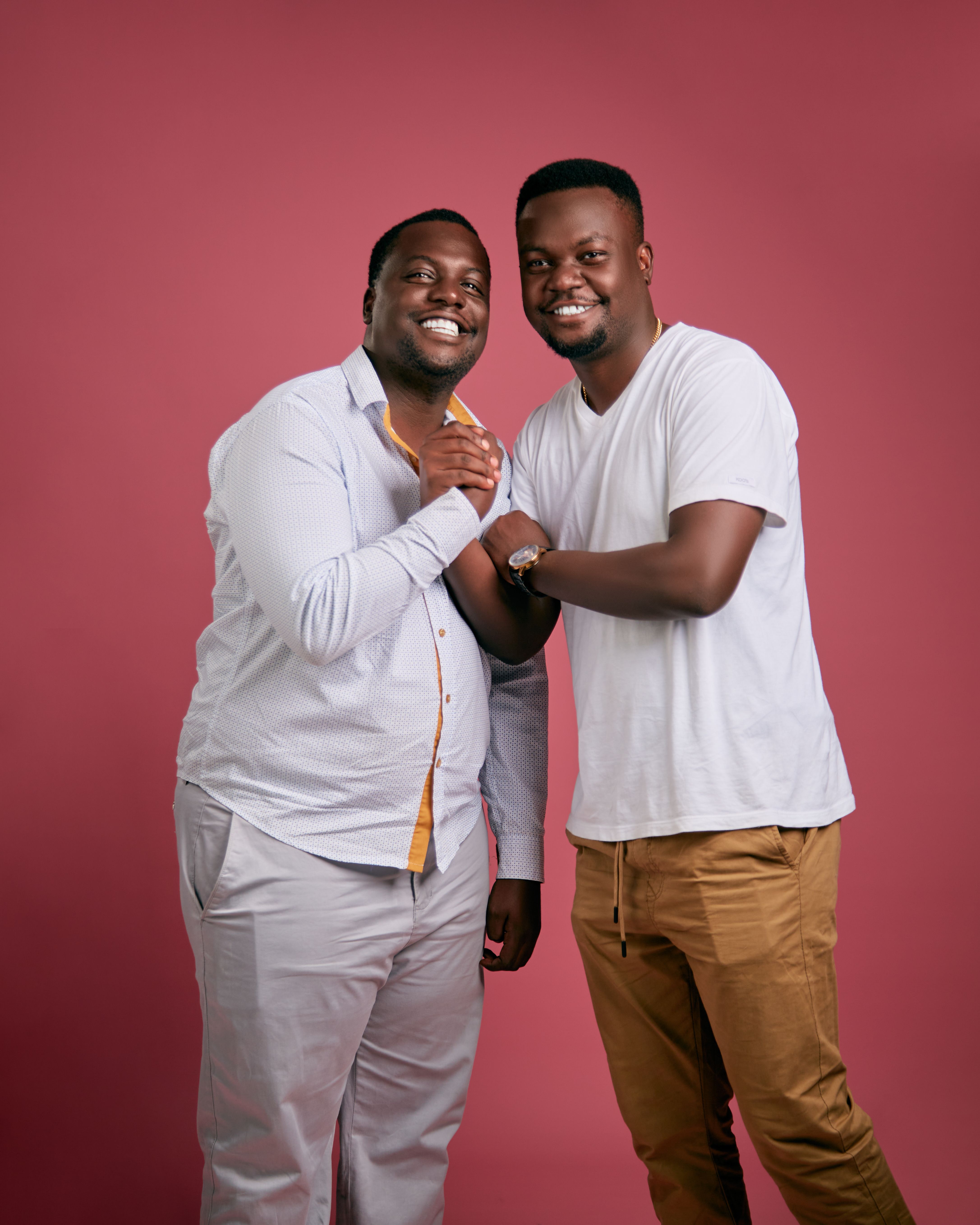 Two men pose happily against a red backdrop, embodying friendship and fashion.