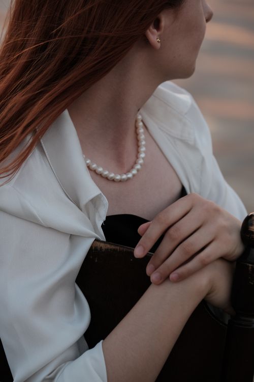 Free A woman with red hair wearing a pearl necklace, posing in soft lighting, partially obscured by a chair. Stock Photo
