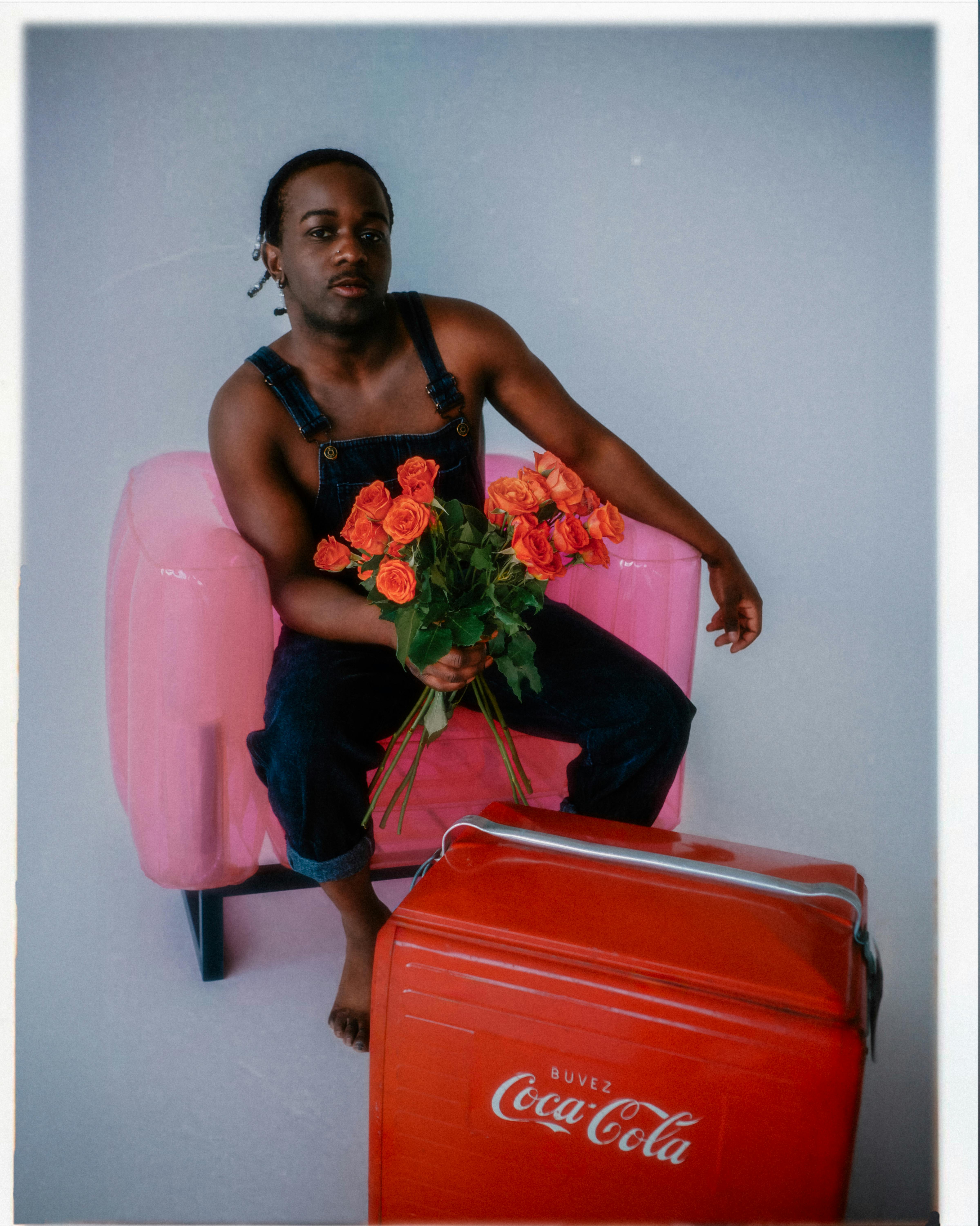 Fashionable studio shoot featuring a shirtless man holding vibrant red roses and sitting on a pink armchair.