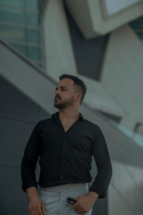 Free Man wearing black shirt and white pants standing in front of modern urban architecture, conveying a sense of style and confidence. Stock Photo