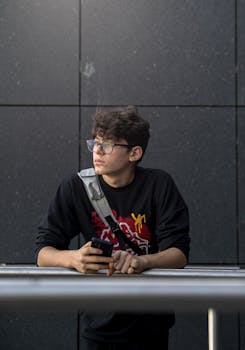 Teenage boy wearing glasses leaning on a metal railing in an urban setting, holding a smartphone.