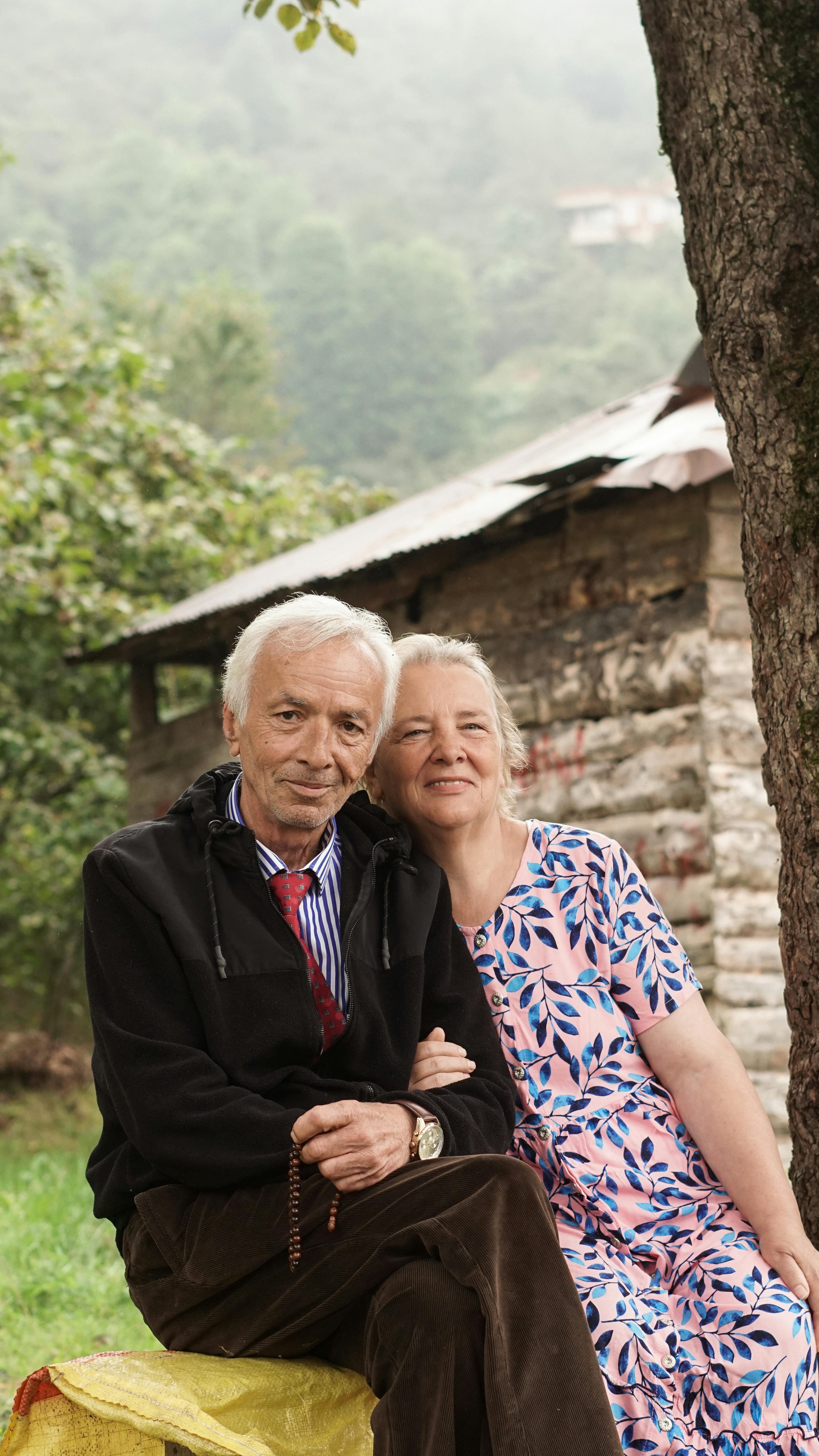 An elderly couple sits together, smiling, in a serene natural setting with rustic architecture.