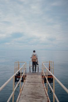 Man stands on a pier with a backpack, looking out at the serene sea.