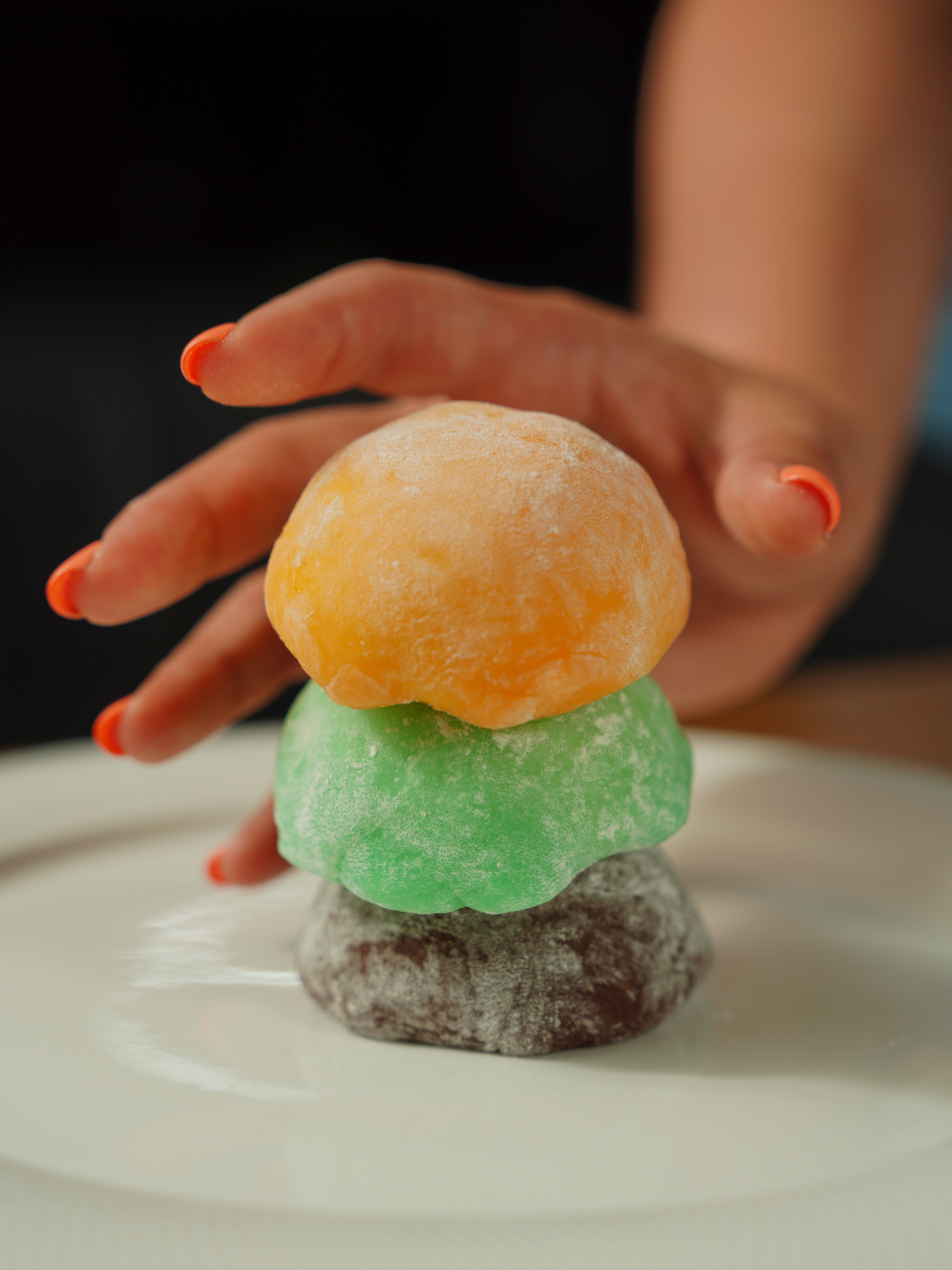 A hand with orange nails reaching for colorful mochi on a plate.