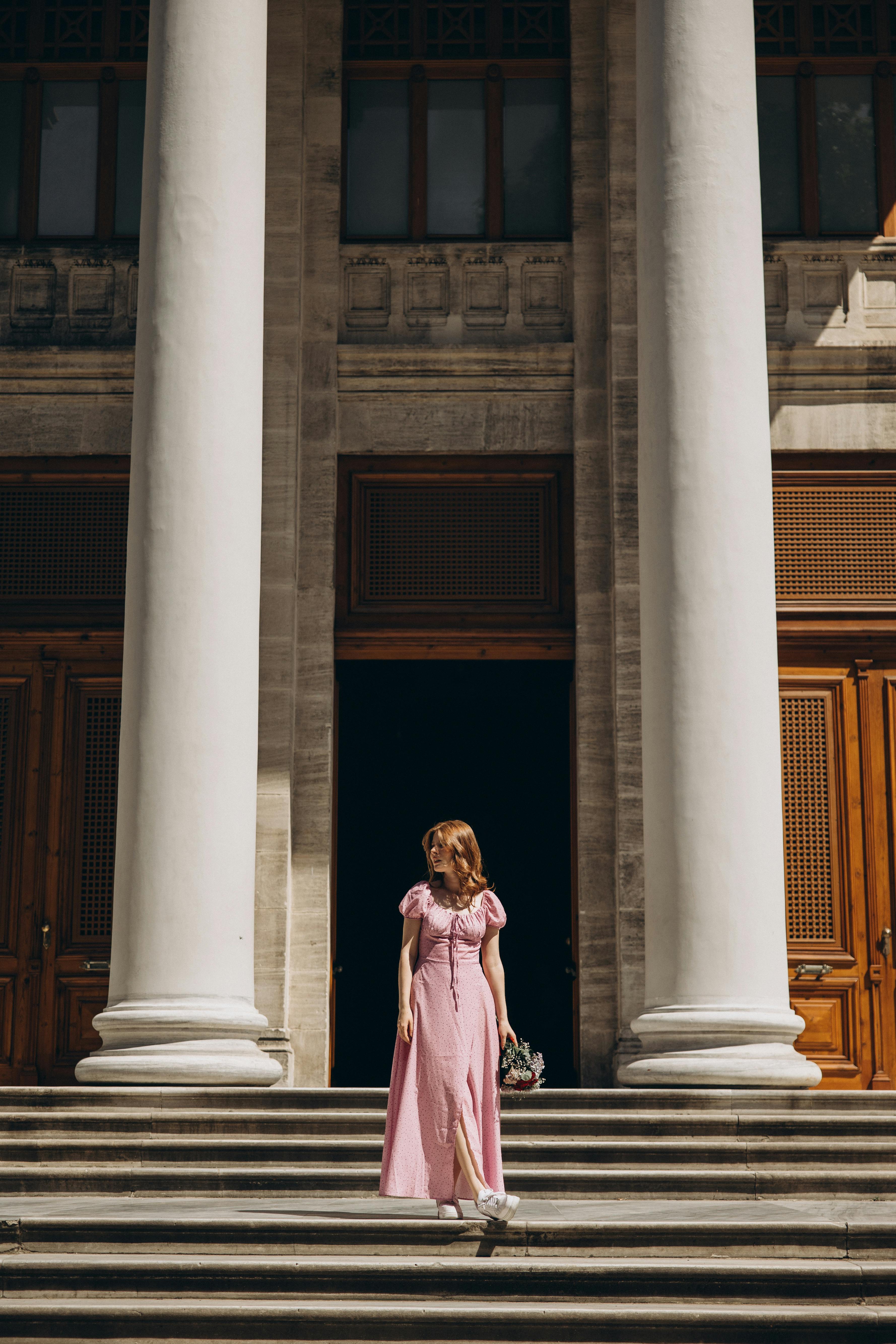 a woman in a pink dress stands on the steps of a building