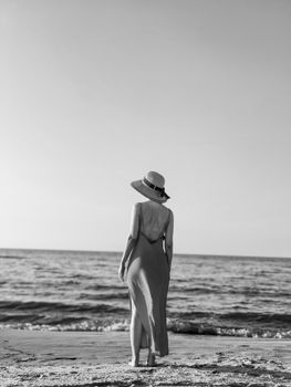 Black and white photo of a woman in a backless dress and straw hat standing on a beach.