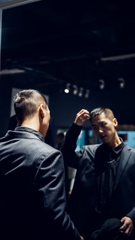 A young man in a dark suit fixing his hair while looking in a mirror indoors.