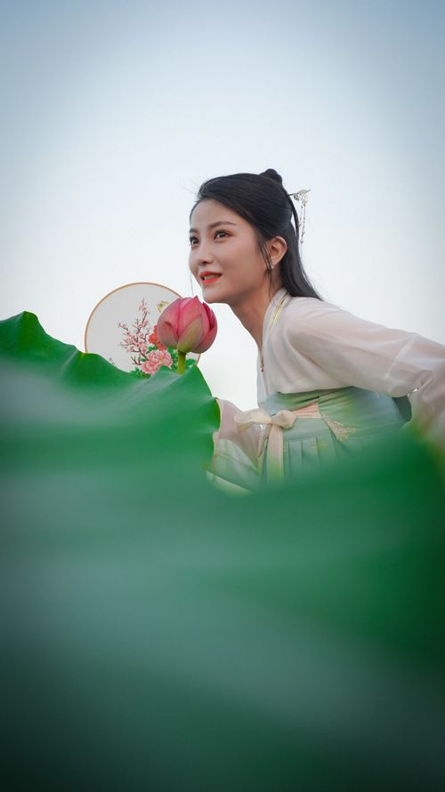 Free Asian woman in traditional attire holding a lotus flower, surrounded by lush leaves, smiling outdoors. Stock Photo