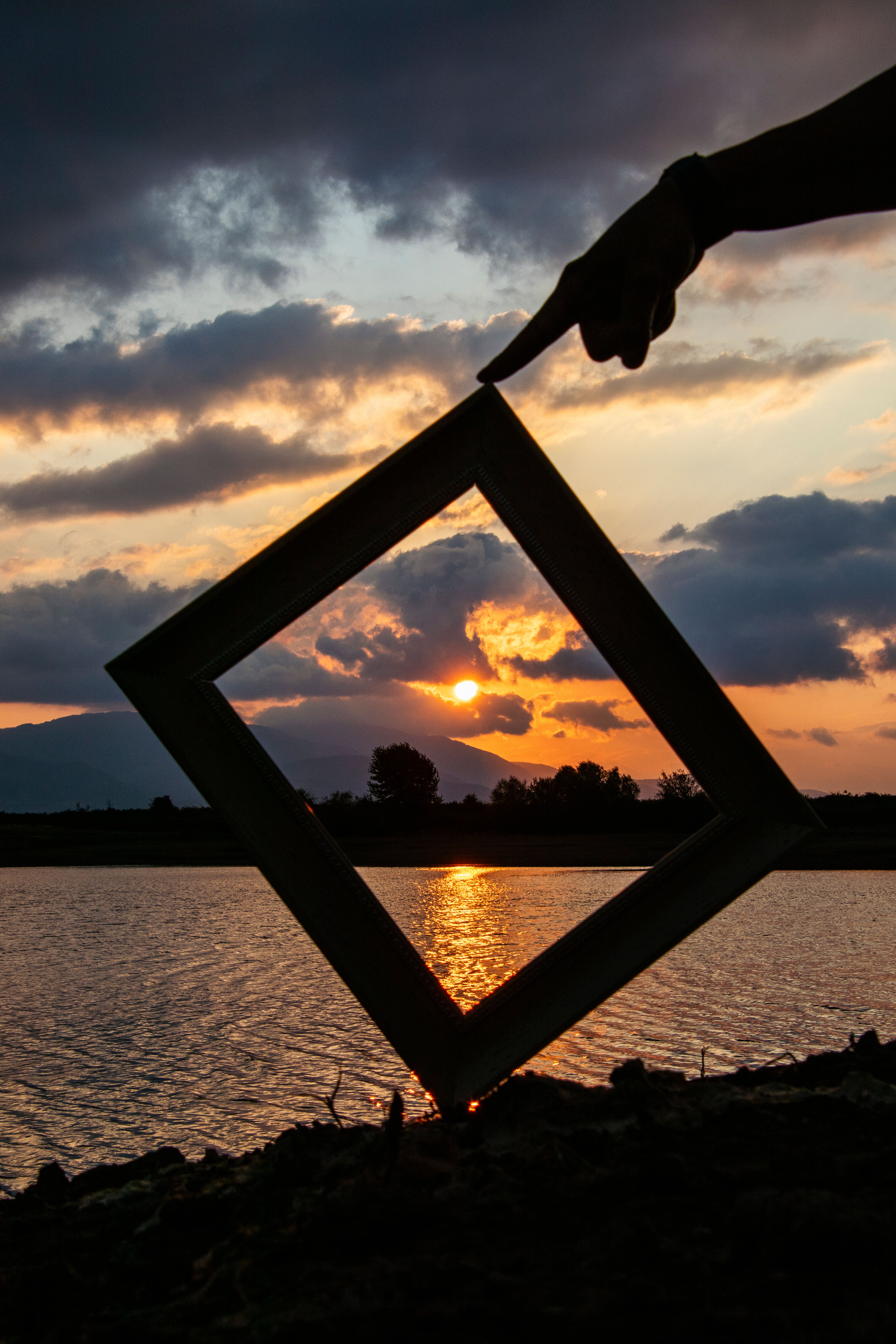 Creative silhouette of a hand holding a frame against a dramatic sunset by the lake.